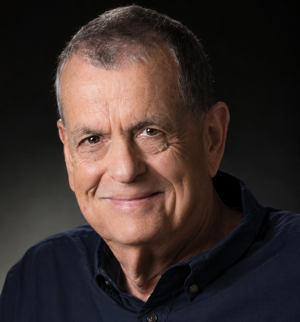 Older man with short gray hair, smiling slightly, wearing a dark shirt against a dark background.